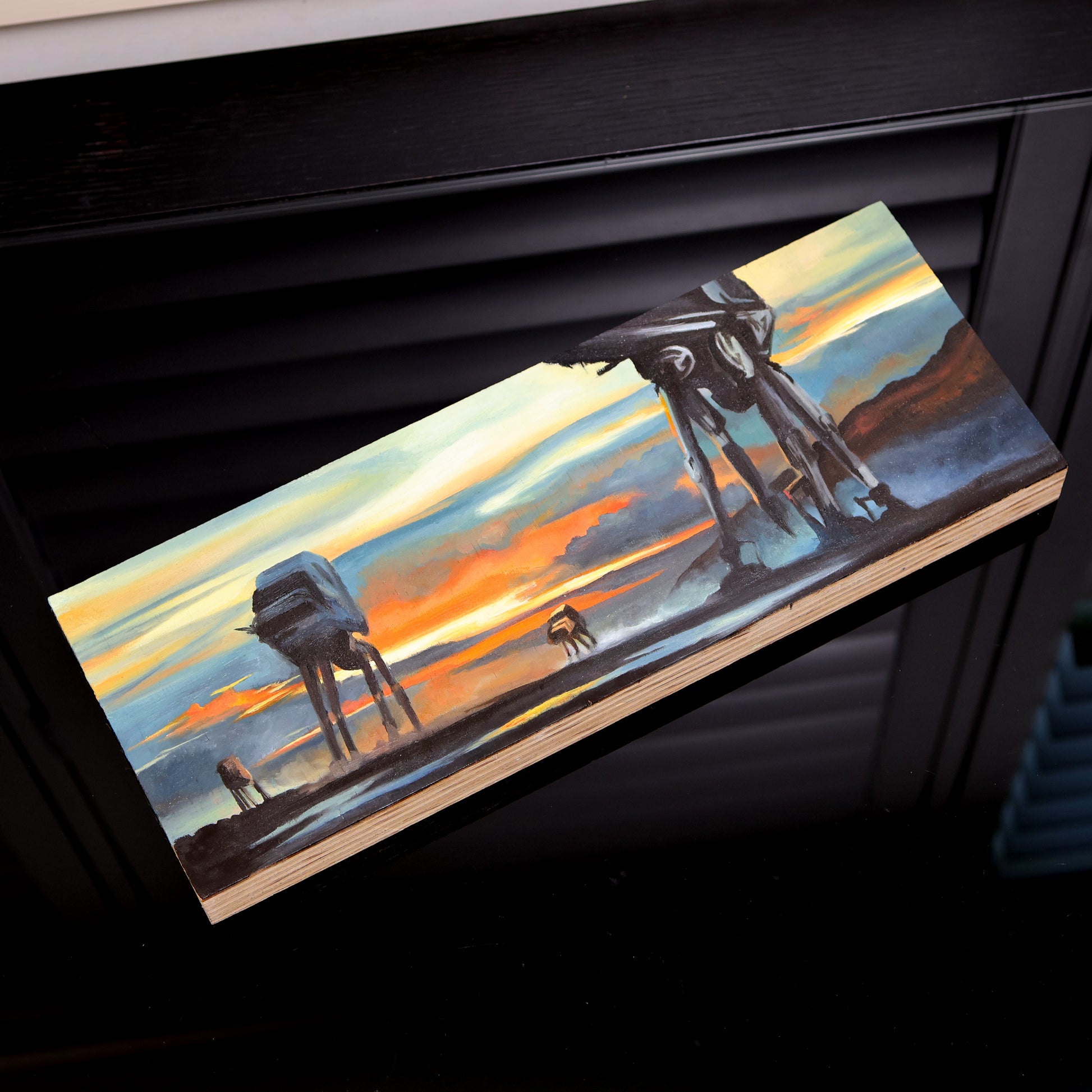 Original oil painting on wood panel depicting silhouetted AT-AT walkers from Star Wars marching across a misty landscape at sunset. Vivid orange, yellow, and blue tones create a dramatic sci-fi atmosphere, with bold contrast and cinematic composition. On black color desk.